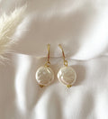 A pair of gold plated Huggies with irregular white baroque pearls, displayed on a soft white fabric background with a decorative beige feather.