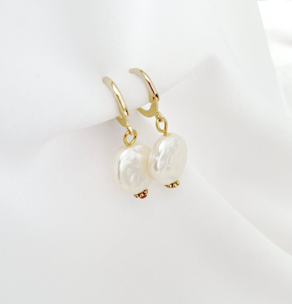 A pair of gold huggie hoop earrings with large, irregular white baroque pearls dangling from the hoops, displayed on a soft white fabric stand.