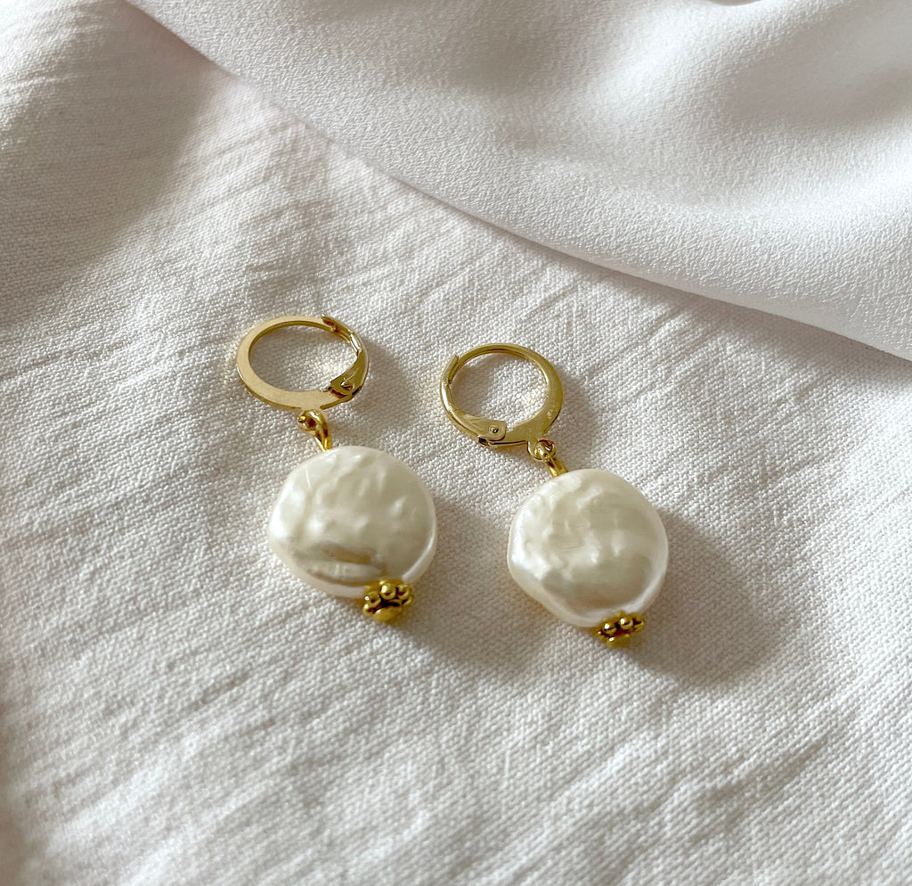 A pair of gold hoop earrings with large, irregular white baroque pearl drops, displayed on a textured white linen fabric.