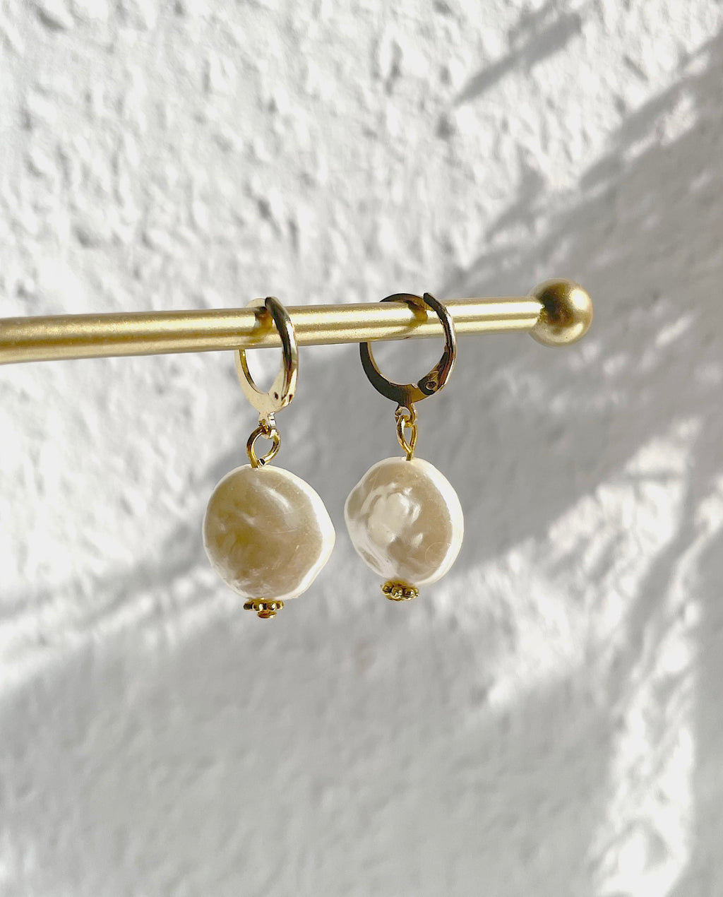 A pair of gold hoop earrings with large, irregular beige colored baroque pearls hanging from a gold rod against a textured white wall with soft leaf shadows.