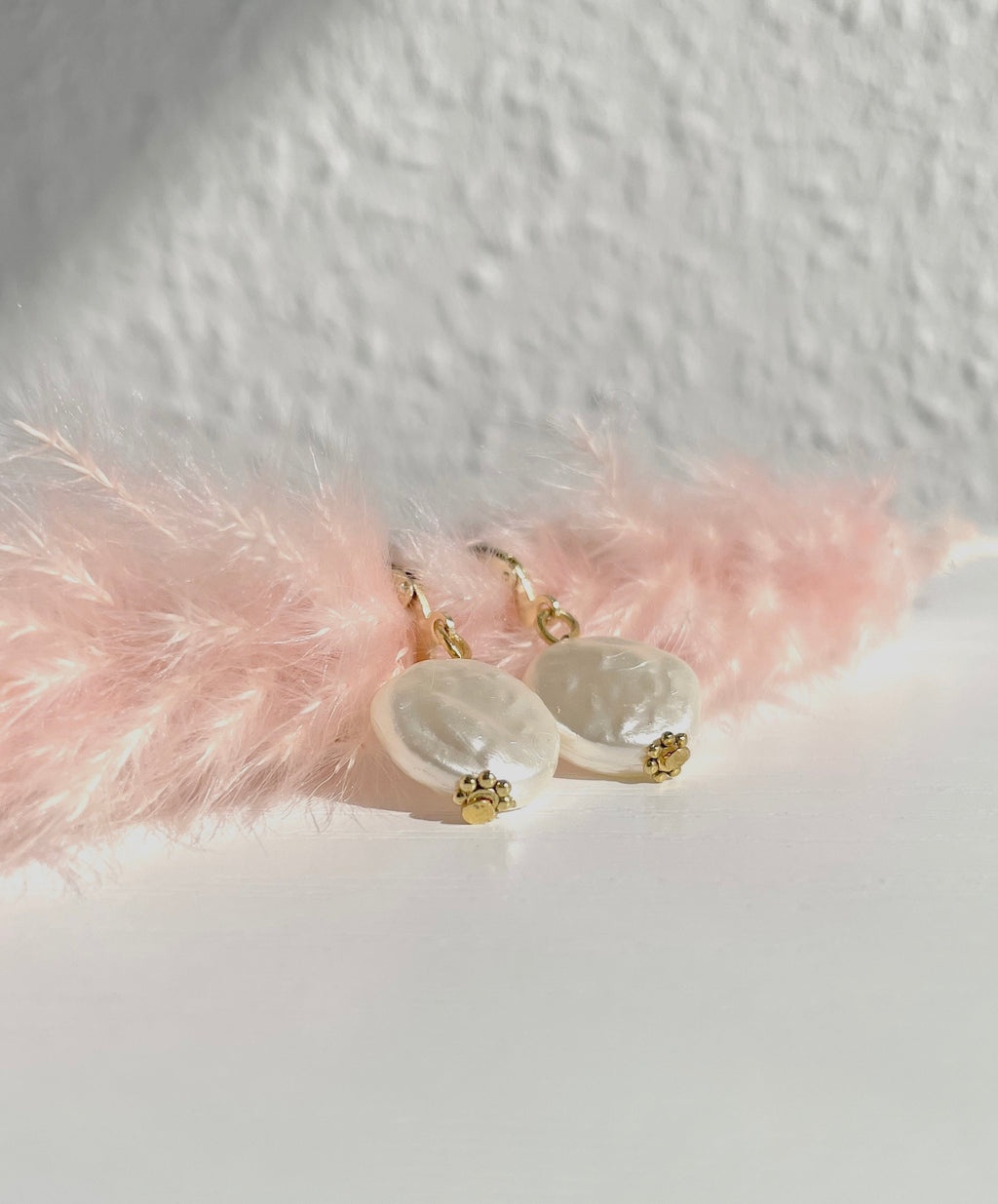 A pair of gold plated pearl dangle earrings with irregular white baroque pearls and small gold floral charms, styled on a white surface with pink decorative feathers against a textured white wall.