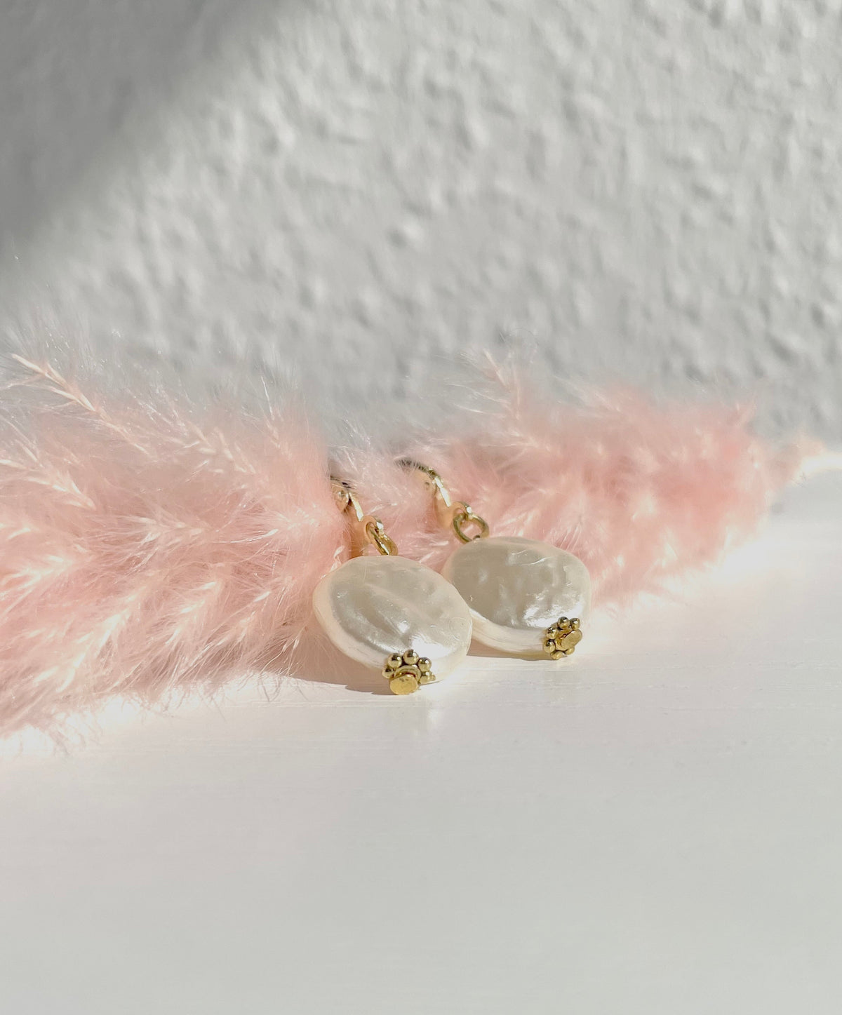 A pair of gold plated pearl dangle earrings with irregular white baroque pearls and small gold floral charms, styled on a white surface with pink decorative feathers against a textured white wall.