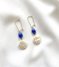 A pair of drop earrings with gold-tone hooks, blue striped beads, and irregular white pearl-like drops, displayed on a white textured fabric background.