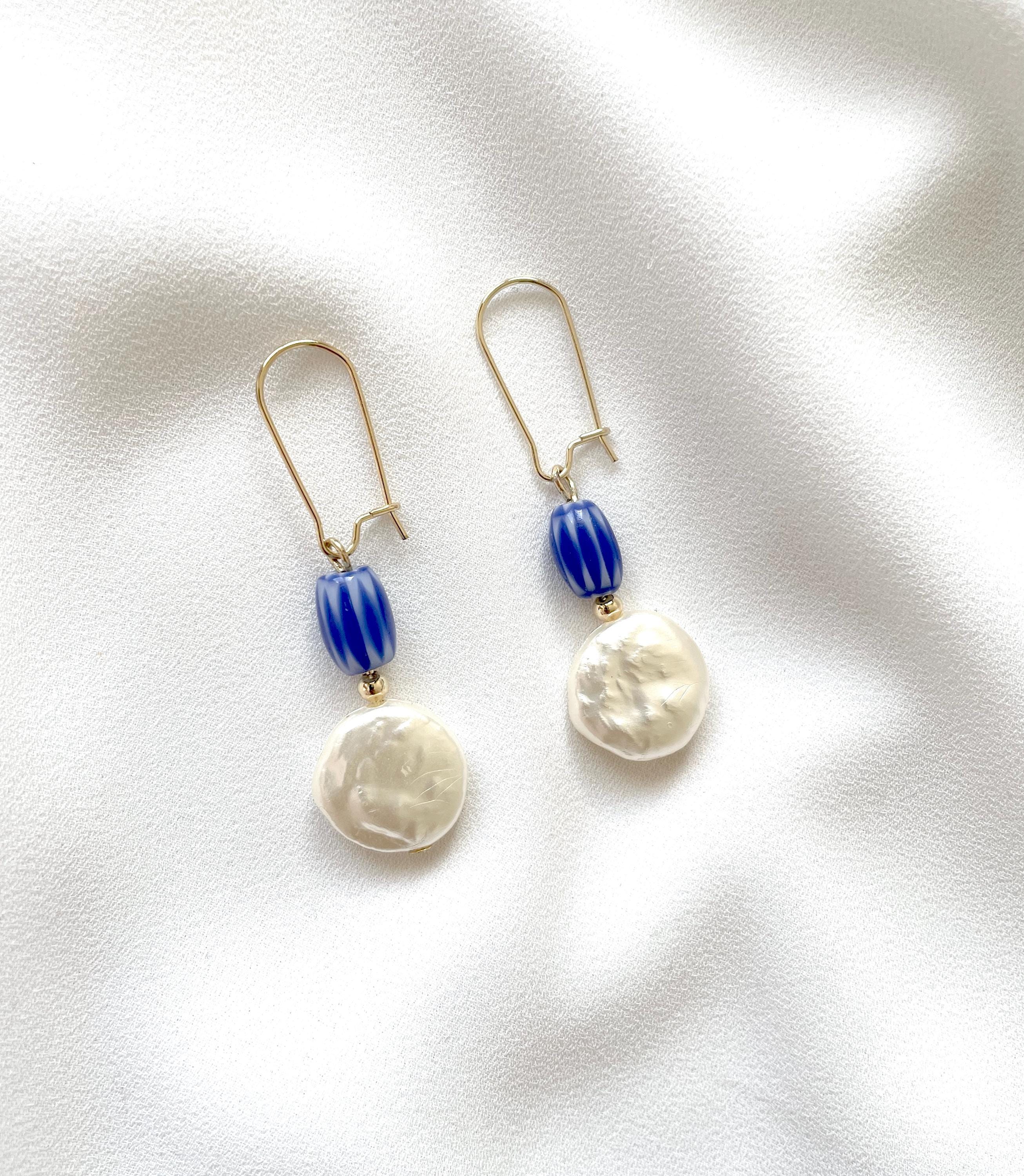 A pair of drop earrings with gold-tone hooks, blue striped beads, and irregular white pearl-like drops, displayed on a white textured fabric background.