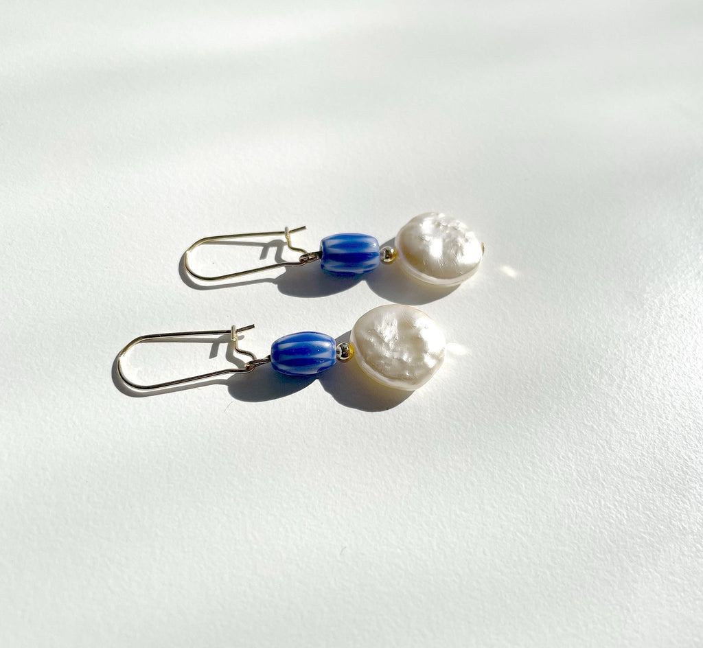 A pair of drop earrings with textured white pearl-like beads, blue striped beads, and silver fishhook wires, displayed on a white surface with soft shadows.