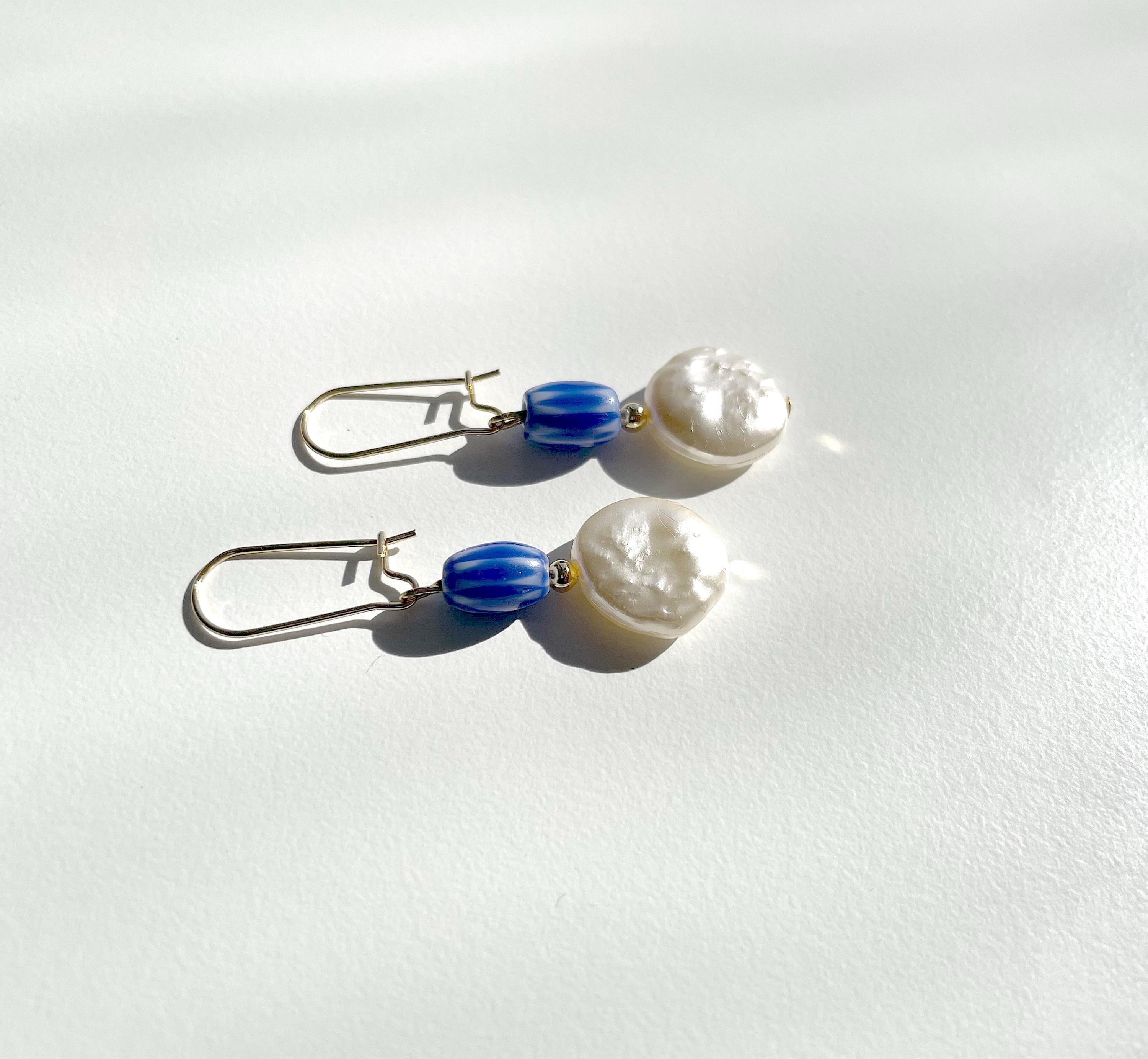 A pair of drop earrings with textured white pearl-like beads, blue striped beads, and silver fishhook wires, displayed on a white surface with soft shadows.