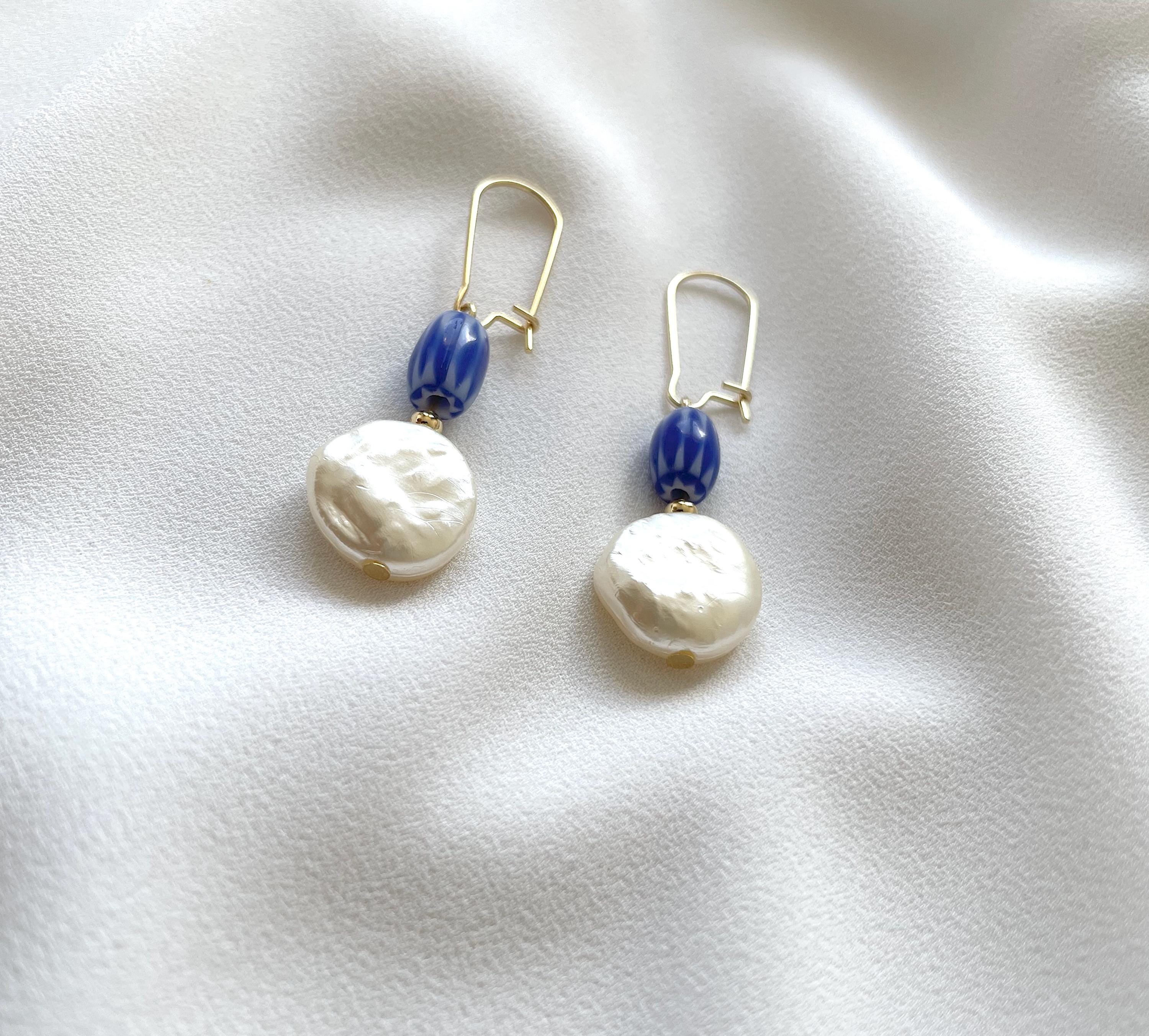 A pair of gold-tone dangle earrings with a small blue striped bead and a large, irregular white baroque pearl, displayed on a soft white fabric background.