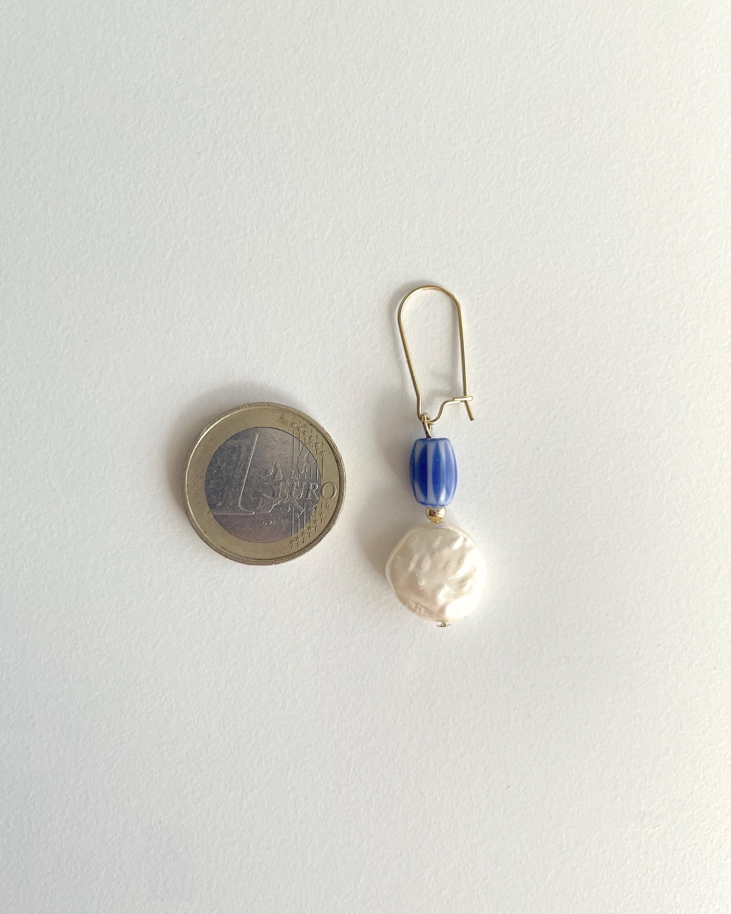 A single drop earring with a gold fishhook back, a small blue faceted bead, and a large irregular white pearl, shown next to a 1 Euro coin for scale on a plain white background.