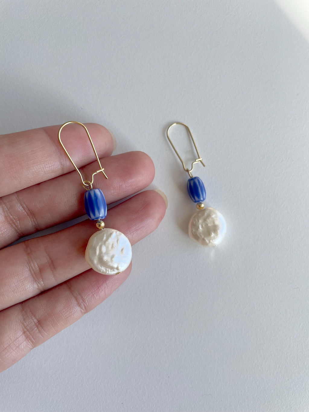 A pair of gold-tone drop earrings with a blue faceted bead and a textured white baroque pearl, shown hanging against a light gray background with a hand holding one earring for scale.