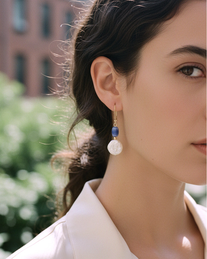 A close-up of a woman wearing a delicate gold dangle earring with a white round pearl bead, a small blue bead, and a tiny gold accent, set against a blurred outdoor background.