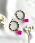 A pair of gold-toned twisted hoop earrings with bright pink bead drops, styled on a white fabric background with small white flowers.