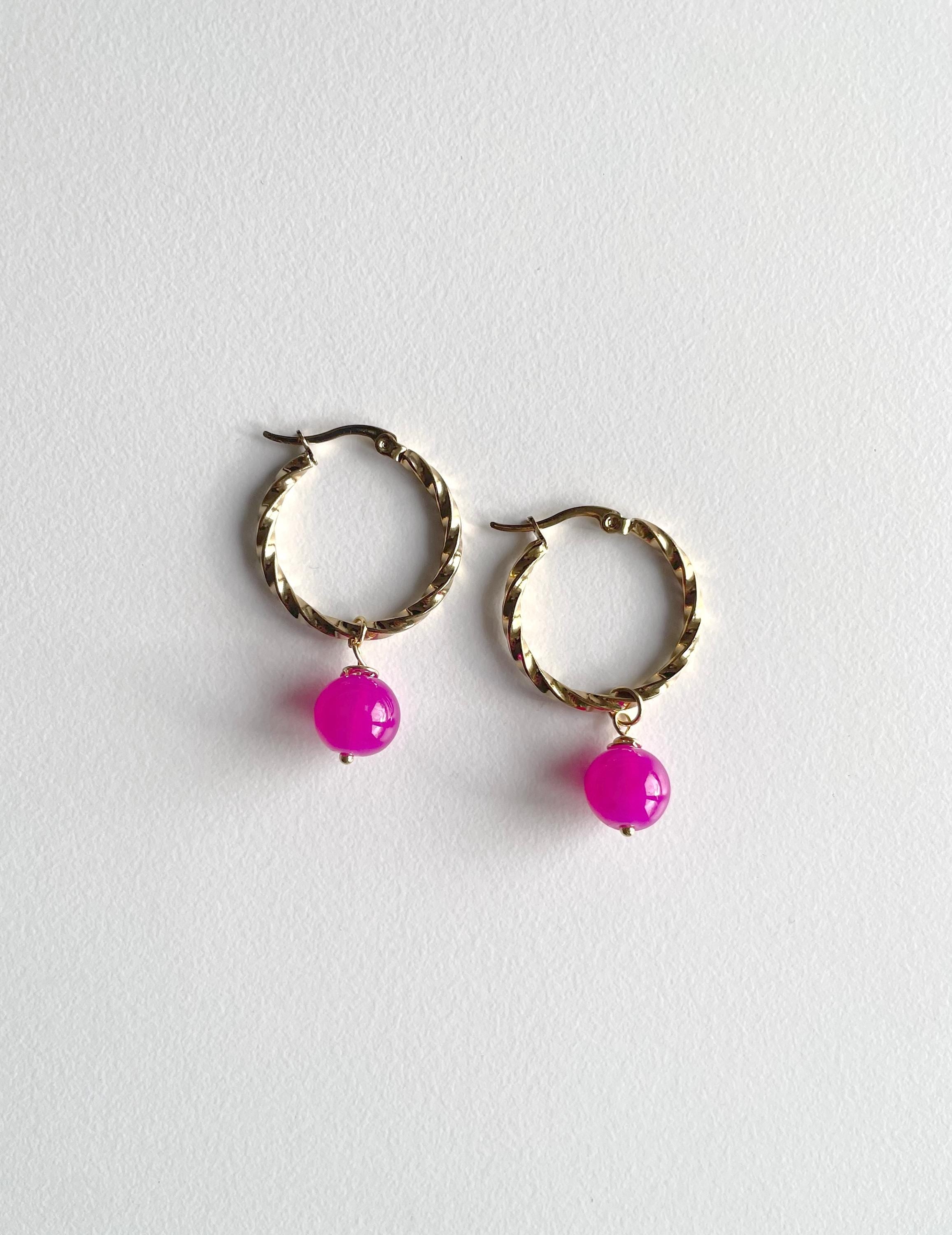 A pair of gold-toned twisted hoop earrings with dangling bright pink bead charms, displayed on a light gray textured background. Hot pink beads. Bright Pink bead, bright pink charms, pink earrings