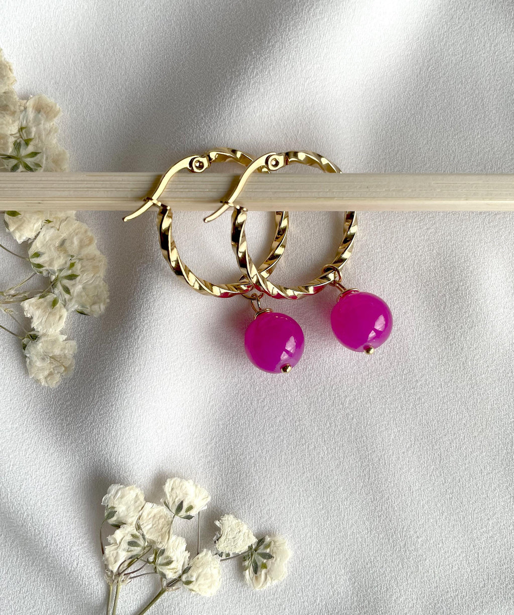 Gold hoop earrings with dangling bright pink bead charms, displayed on a light wooden stick against a white fabric background with small white flowers.