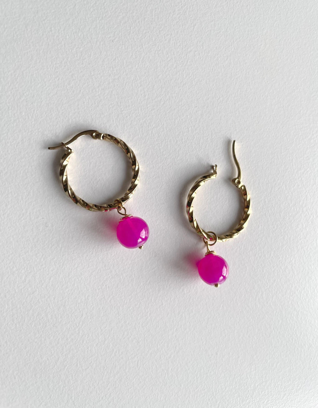 A pair of gold-toned twisted hoop earrings with dangling bright pink faceted beads, displayed on a light gray textured background.