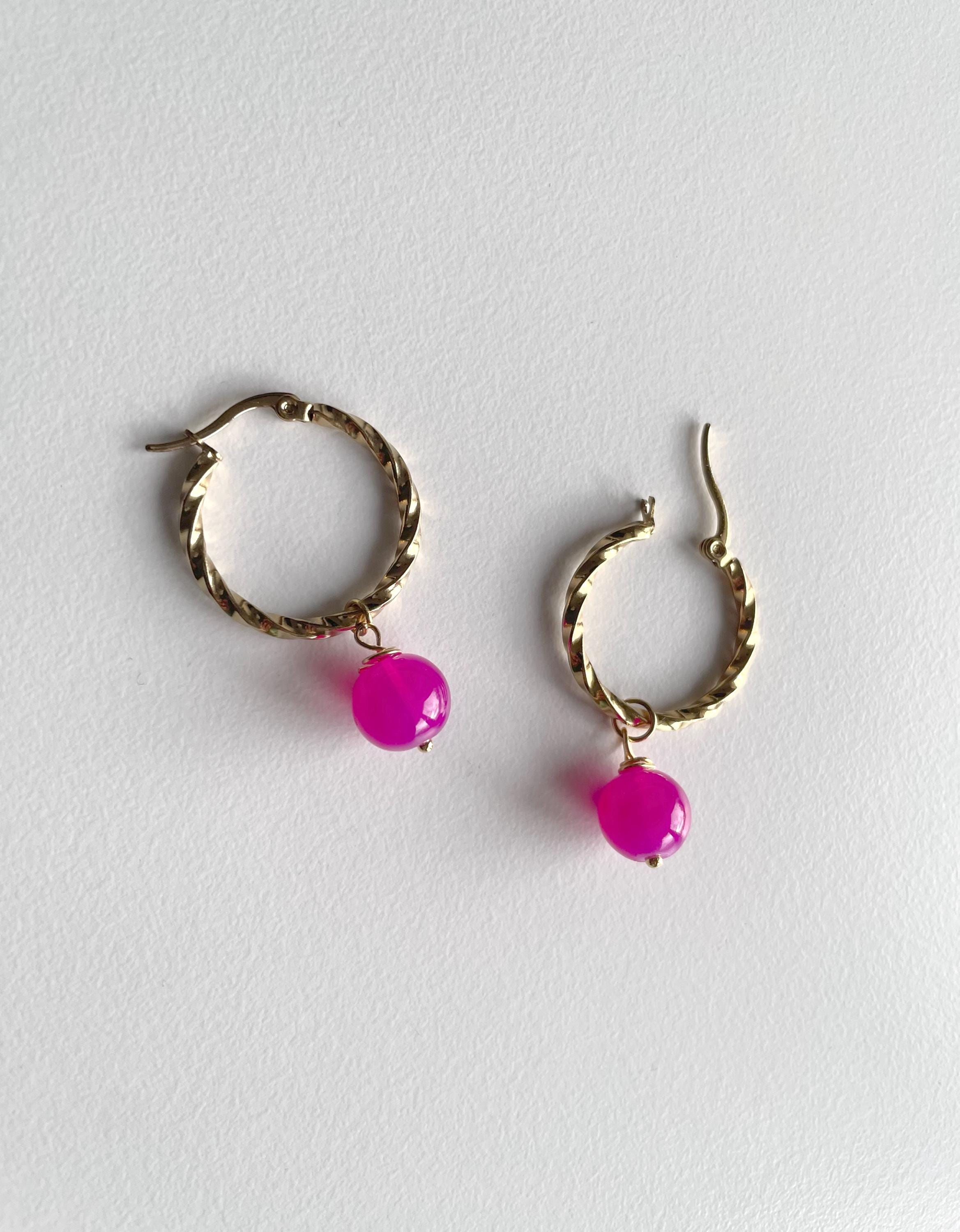 A pair of gold-toned twisted hoop earrings with dangling bright pink faceted beads, displayed on a light gray textured background.