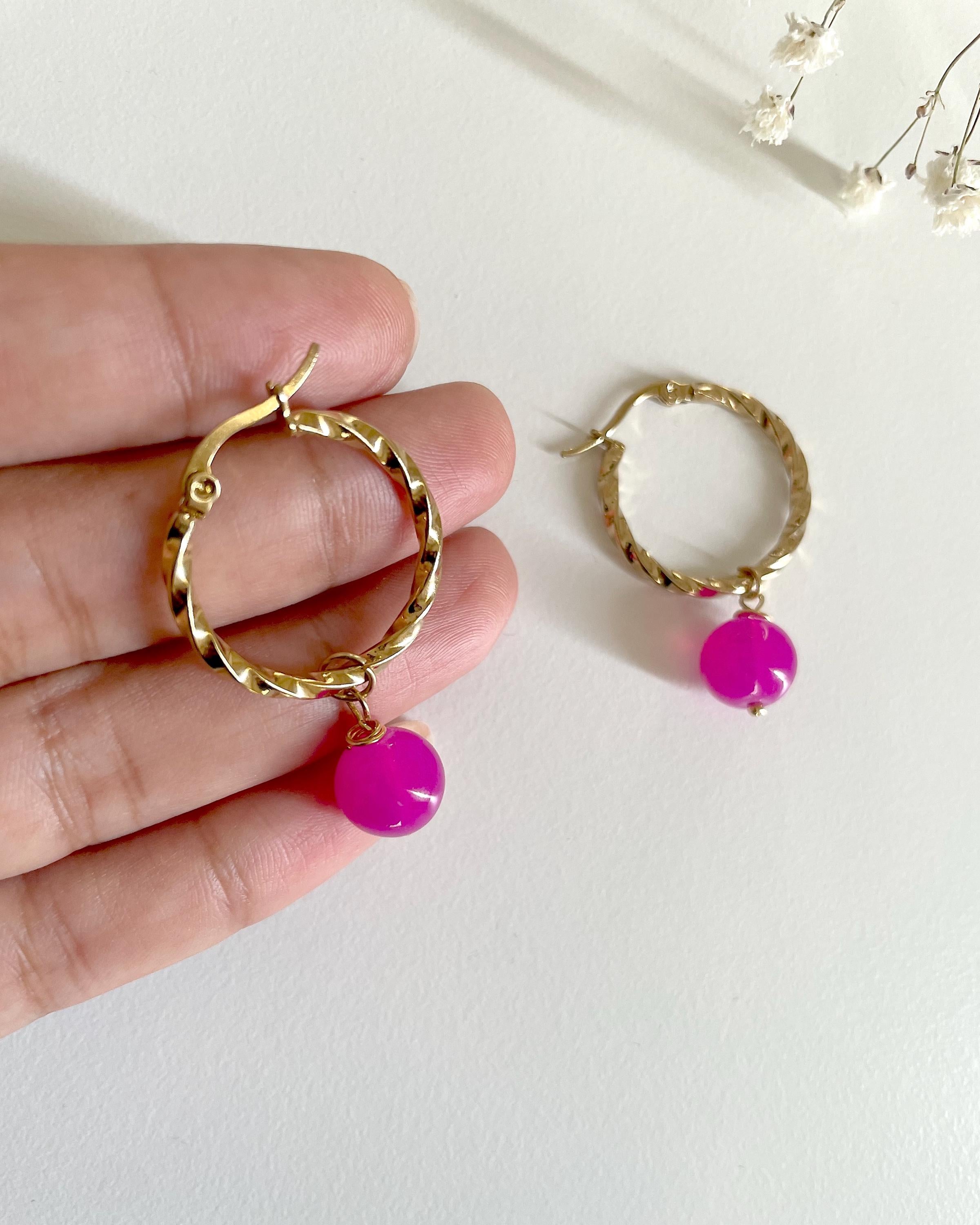Gold hoop earrings with dangling bright pink beads, shown with one held on a person's hand and one resting on a white surface.