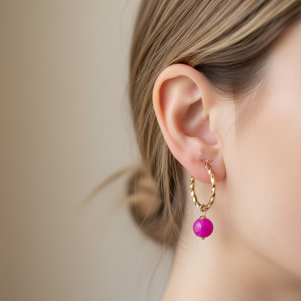 A close-up of a person's ear wearing a gold hoop earring with a dangling bright pink bead.