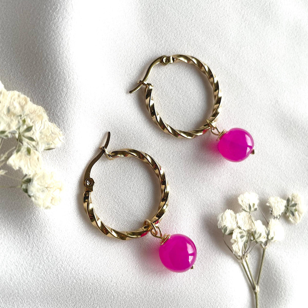 A pair of gold-toned twisted hoop earrings with bright pink bead drops, styled on a white fabric background with small white flowers.