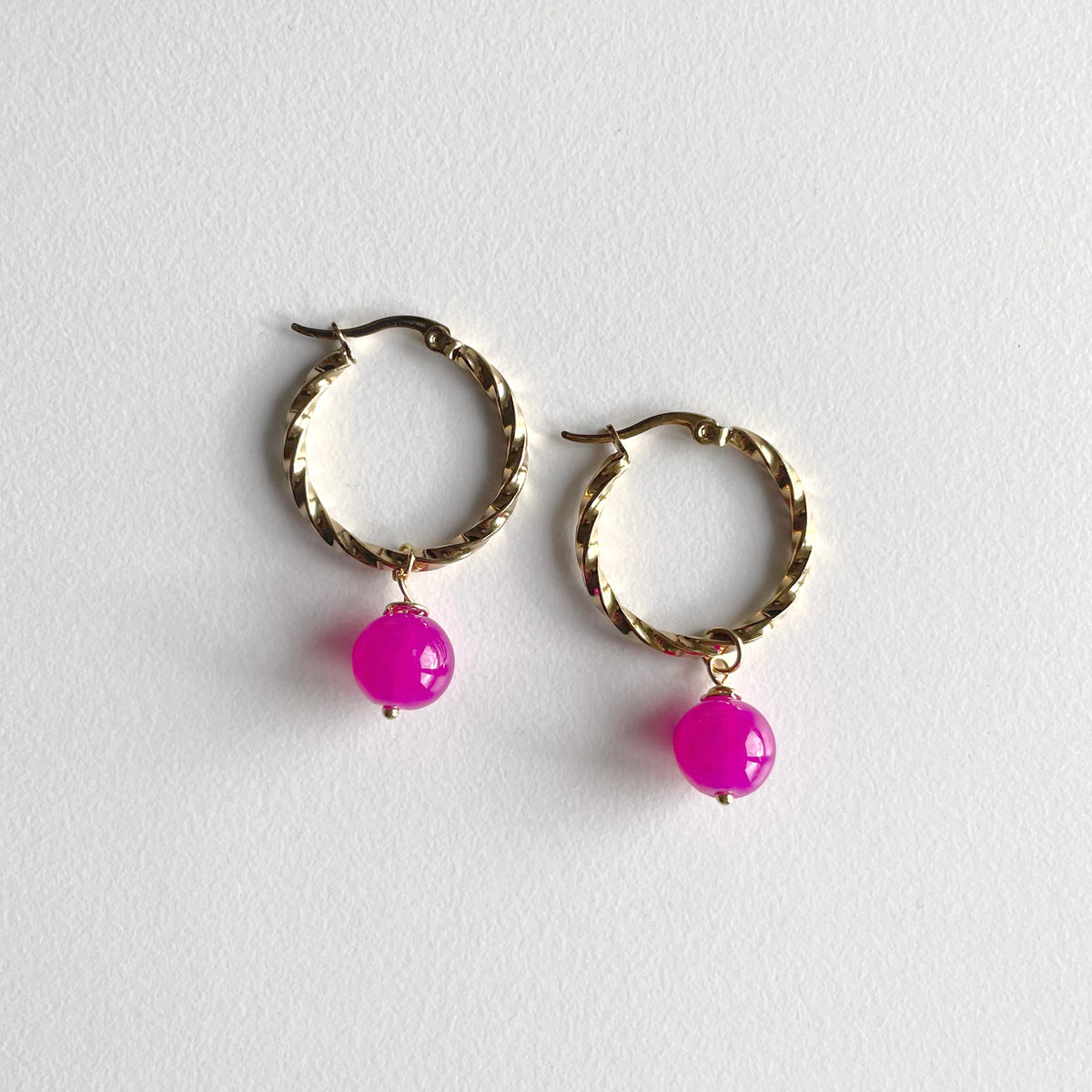 A pair of gold-toned twisted hoop earrings with dangling bright pink bead charms, displayed on a light gray textured background. Hot pink beads. Bright Pink bead, bright pink charms, pink earrings