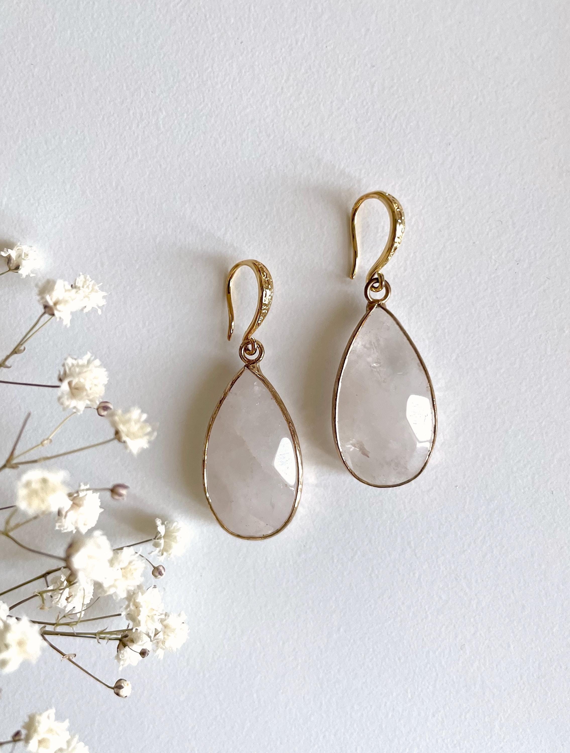 A pair of gold drop earrings with teardrop-shaped clear faceted stones, displayed on a light textured background next to small white flowers.