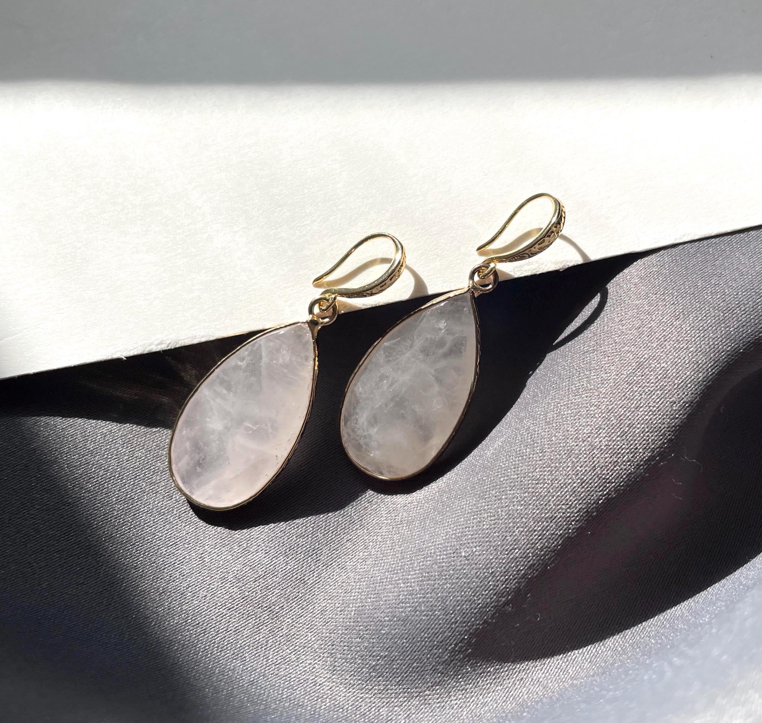 A pair of gold drop earrings with teardrop-shaped pale pinkish-white stone pendants, resting on a dark gray textured surface with strong sunlight and shadows.