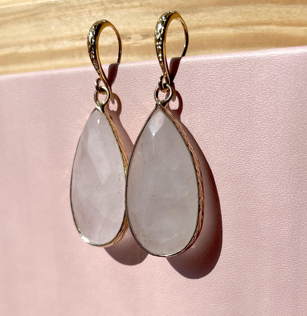 A pair of gold drop earrings with teardrop-shaped pale pink stone pendants, displayed against a soft pink background with a light wood surface at the top.