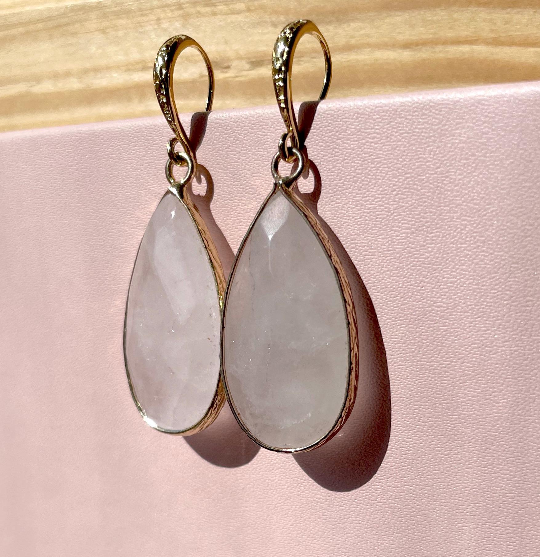 A pair of gold drop earrings with teardrop-shaped pale pink stone pendants, displayed against a soft pink background with a light wood surface at the top.
