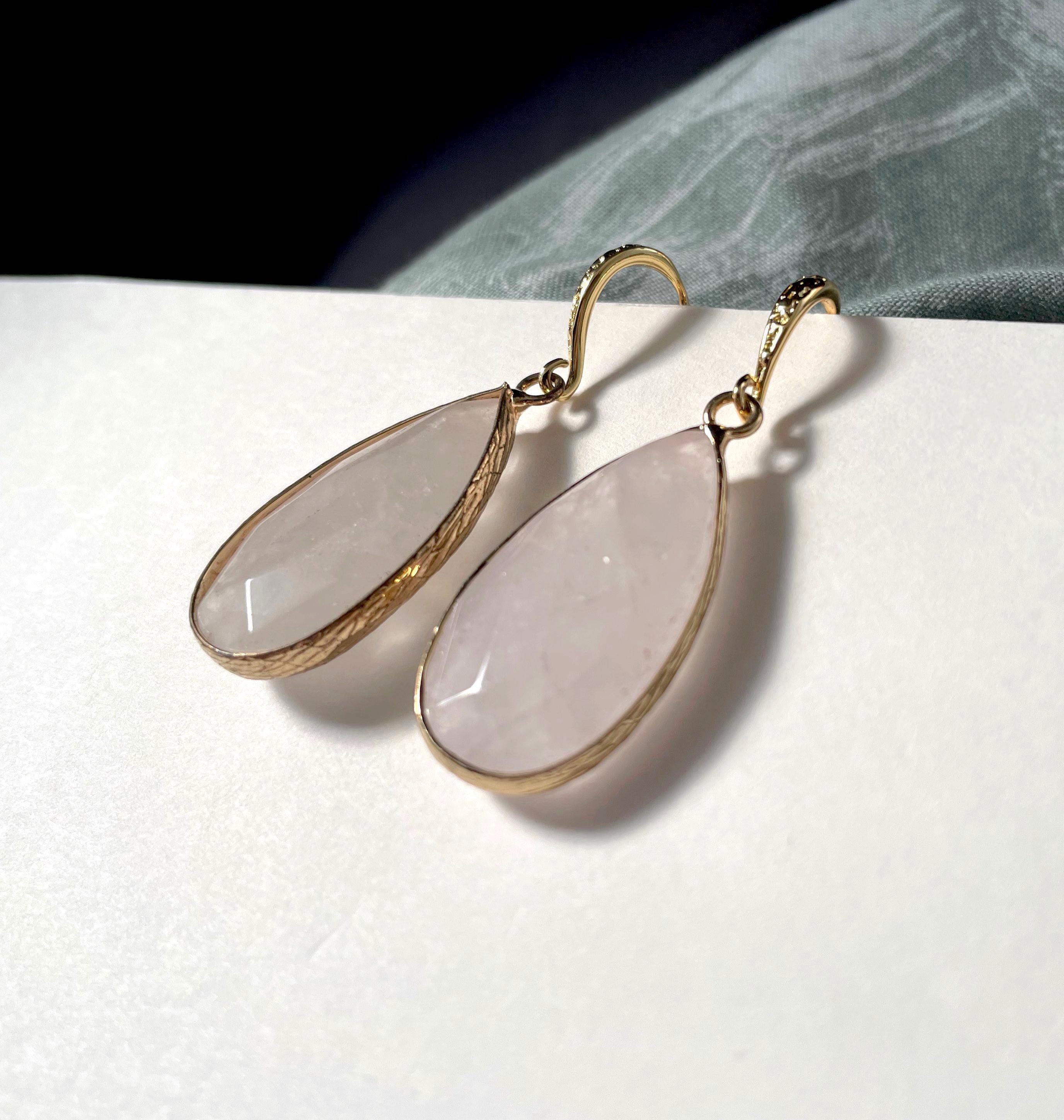 A pair of gold drop earrings with teardrop-shaped pale pink stones, resting on a white textured surface with a dark background and a gray fabric accent.