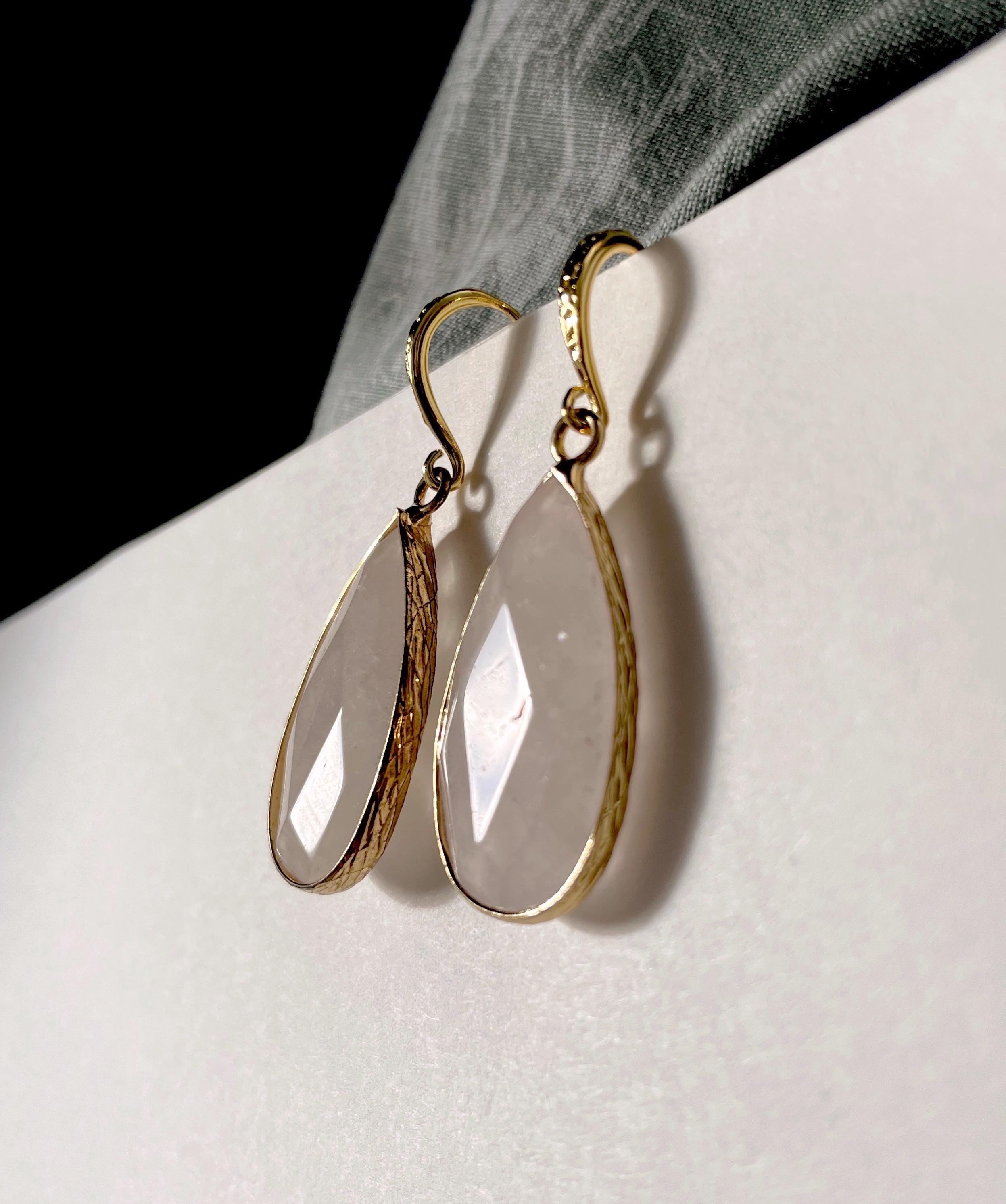 A pair of gold drop earrings with teardrop-shaped pale pinkish-white stones, displayed on a white surface with a dark gray fabric accent.