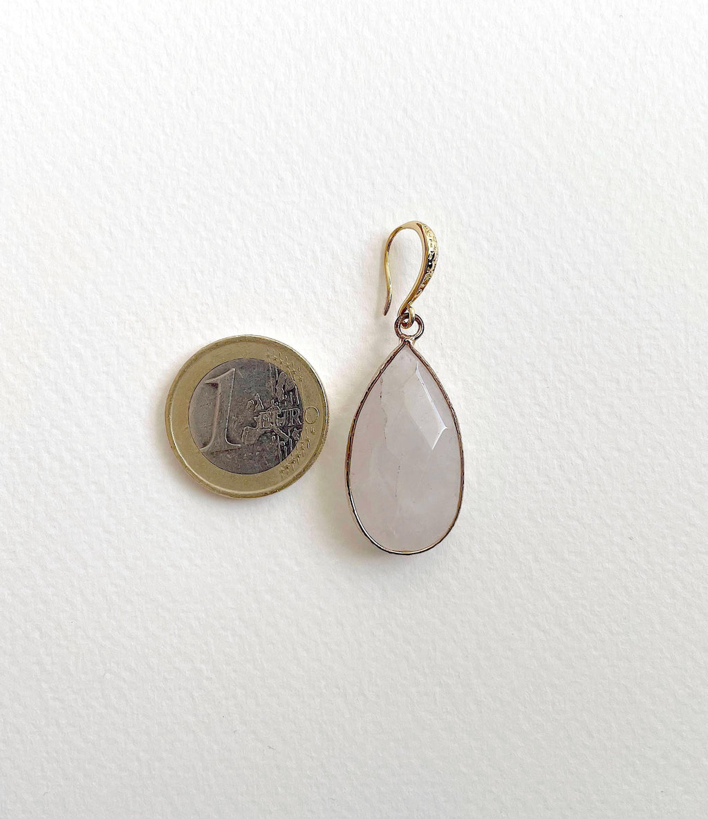 A teardrop-shaped pendant with a pale pink stone in a gold-toned setting, shown next to a 1 euro coin for scale on a white textured background.