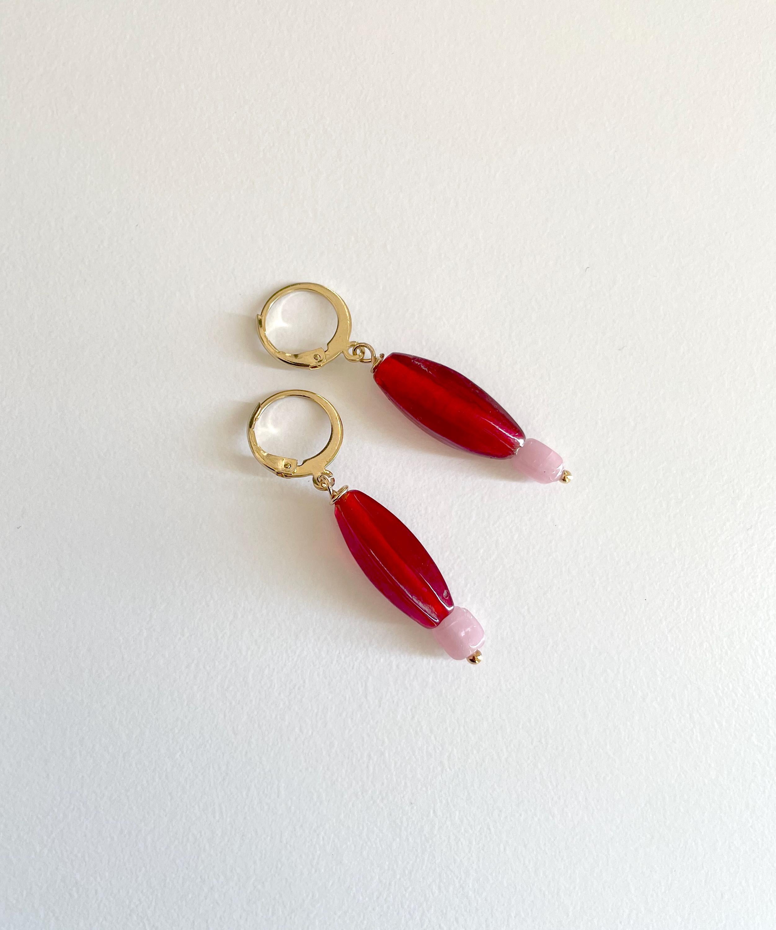 A pair of gold hoop earrings with elongated red beads and small pale pink tips, photographed on a plain white background.