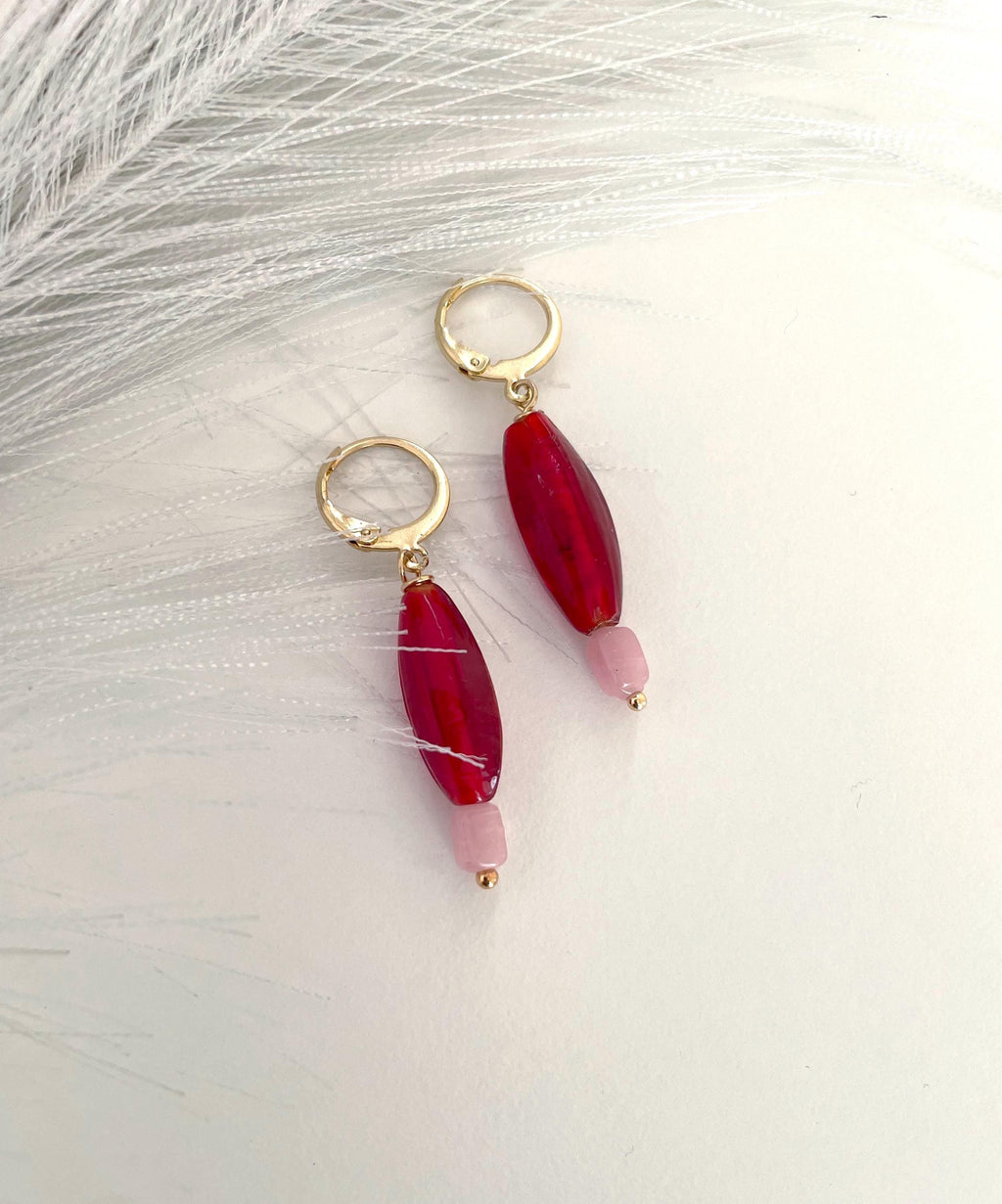 A pair of gold hoop drop earrings featuring a large, faceted deep red bead and a smaller light pink bead, displayed on a soft white textured surface.