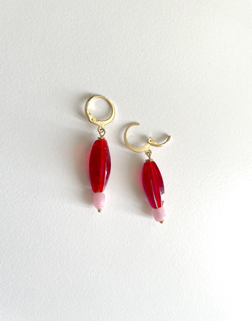 A pair of gold hoop earrings with dangling red teardrop beads and light pink tips, displayed on a clean white background.