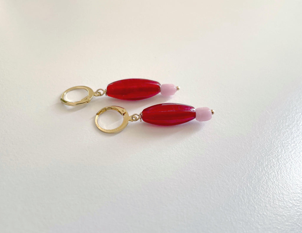 A pair of gold hoop earrings with elongated red beads and small light pink beads, displayed on a plain white surface.