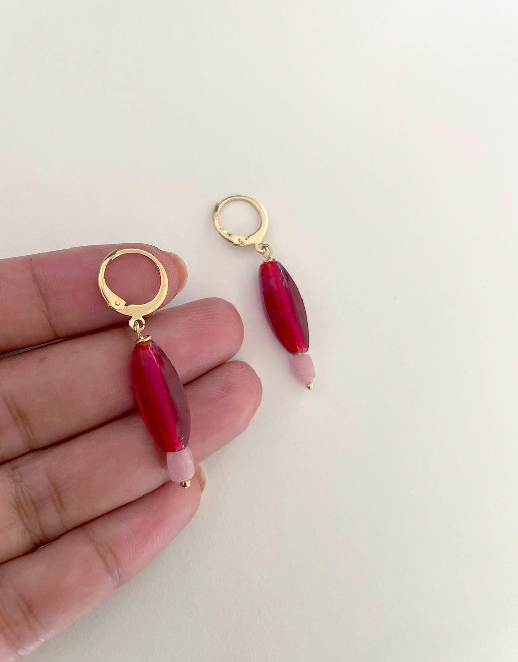 A pair of gold hoop earrings with faceted red beads and light pink tips, shown with one earring hanging and the other held in a hand for scale against a plain off-white background.