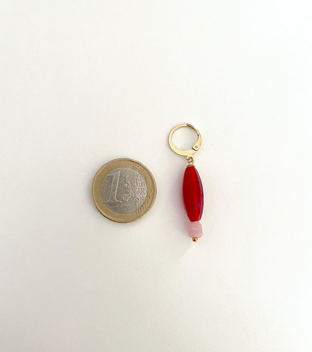 A small gold hoop earring with a dangling red bead and pale pink accent bead, shown next to a 1 Euro coin for scale on a plain white background.