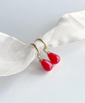 A pair of gold hoop earrings with bright red teardrop-shaped drop charms, displayed on a white satin ribbon against a light gray background.