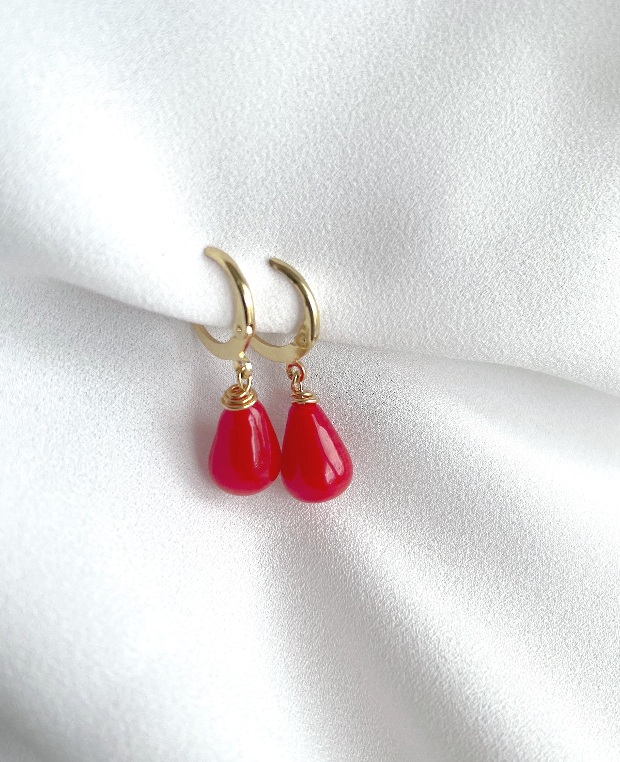 A pair of gold huggie hoop earrings with dangling red teardrop beads, displayed on a soft, white textured fabric background.