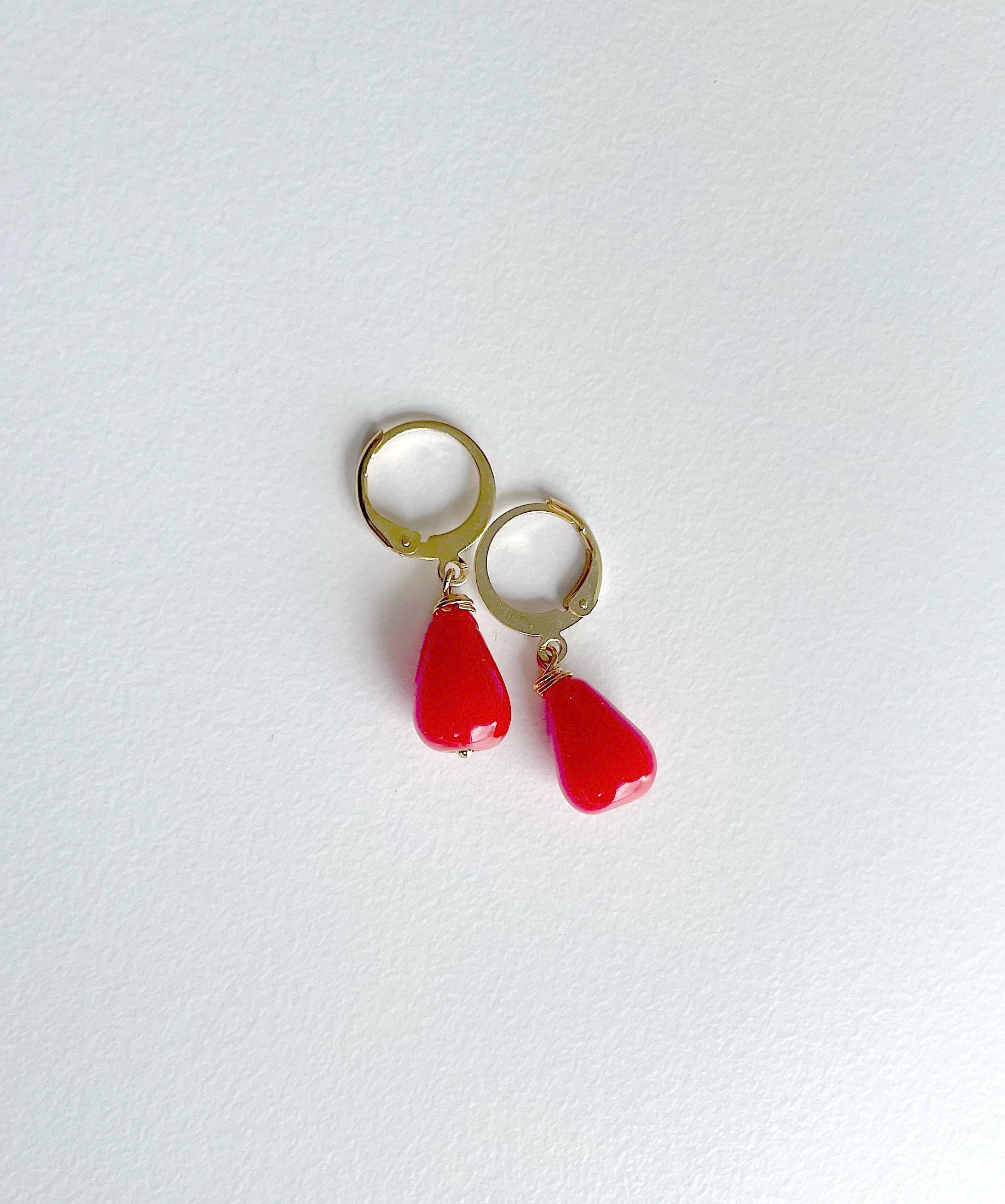 A pair of gold hoop earrings with dangling red teardrop-shaped beads, displayed on a clean, light off-white textured surface.