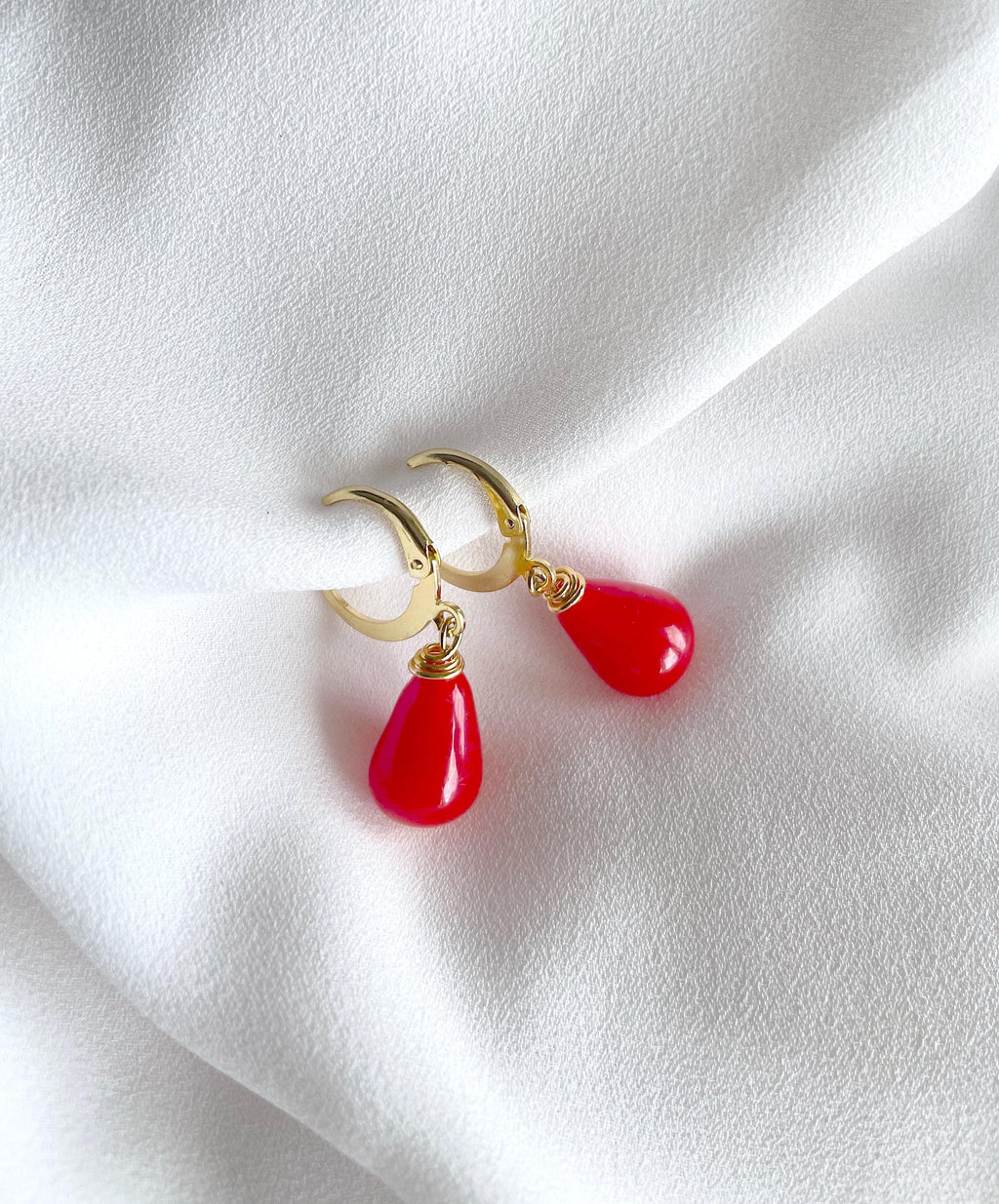 A pair of gold hoop earrings with glossy red teardrop drop charms, displayed on a draped white fabric background.