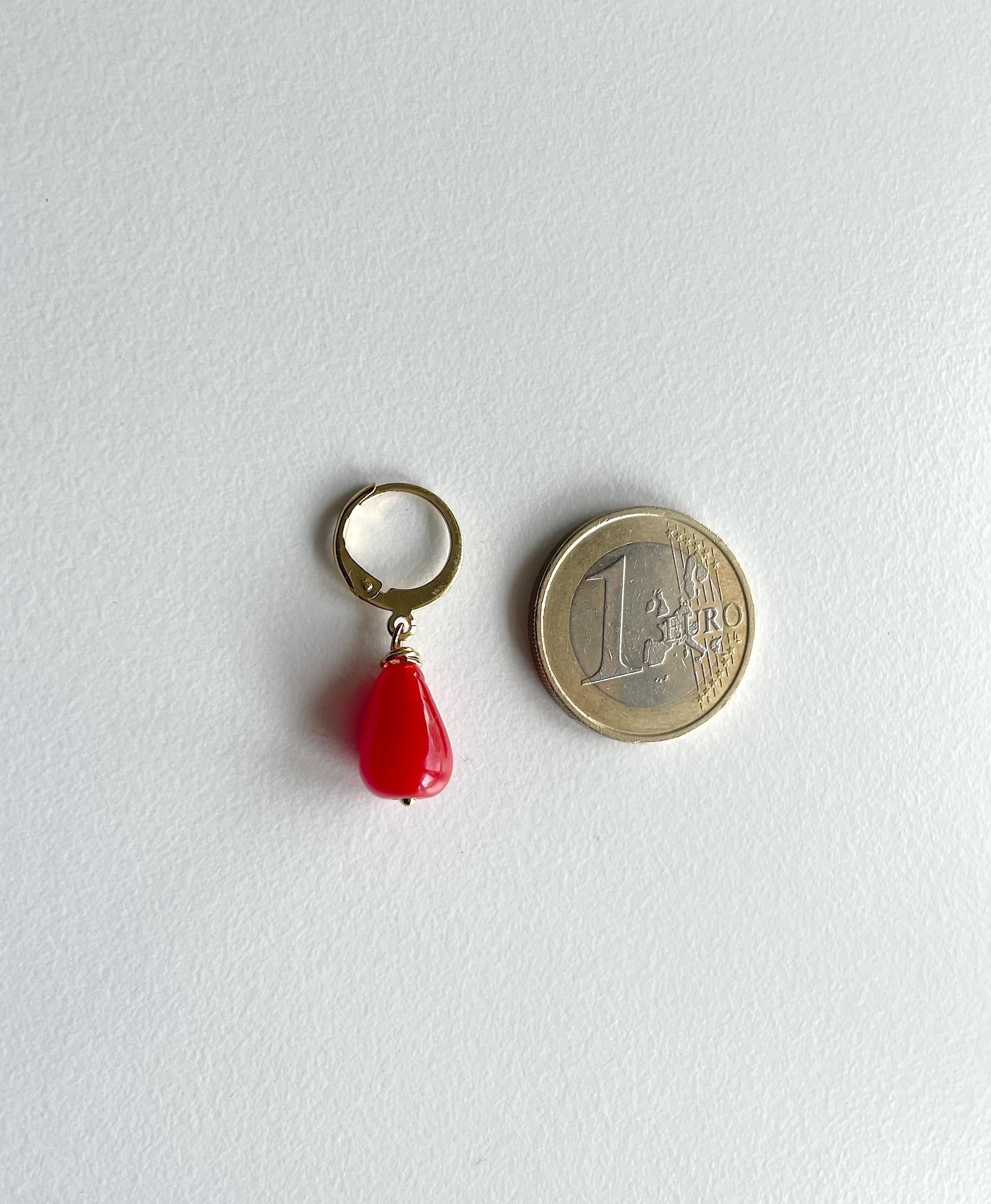 A small gold-tone ring with a dangling red teardrop bead charm, shown next to a 1 euro coin for scale on a light gray textured background.