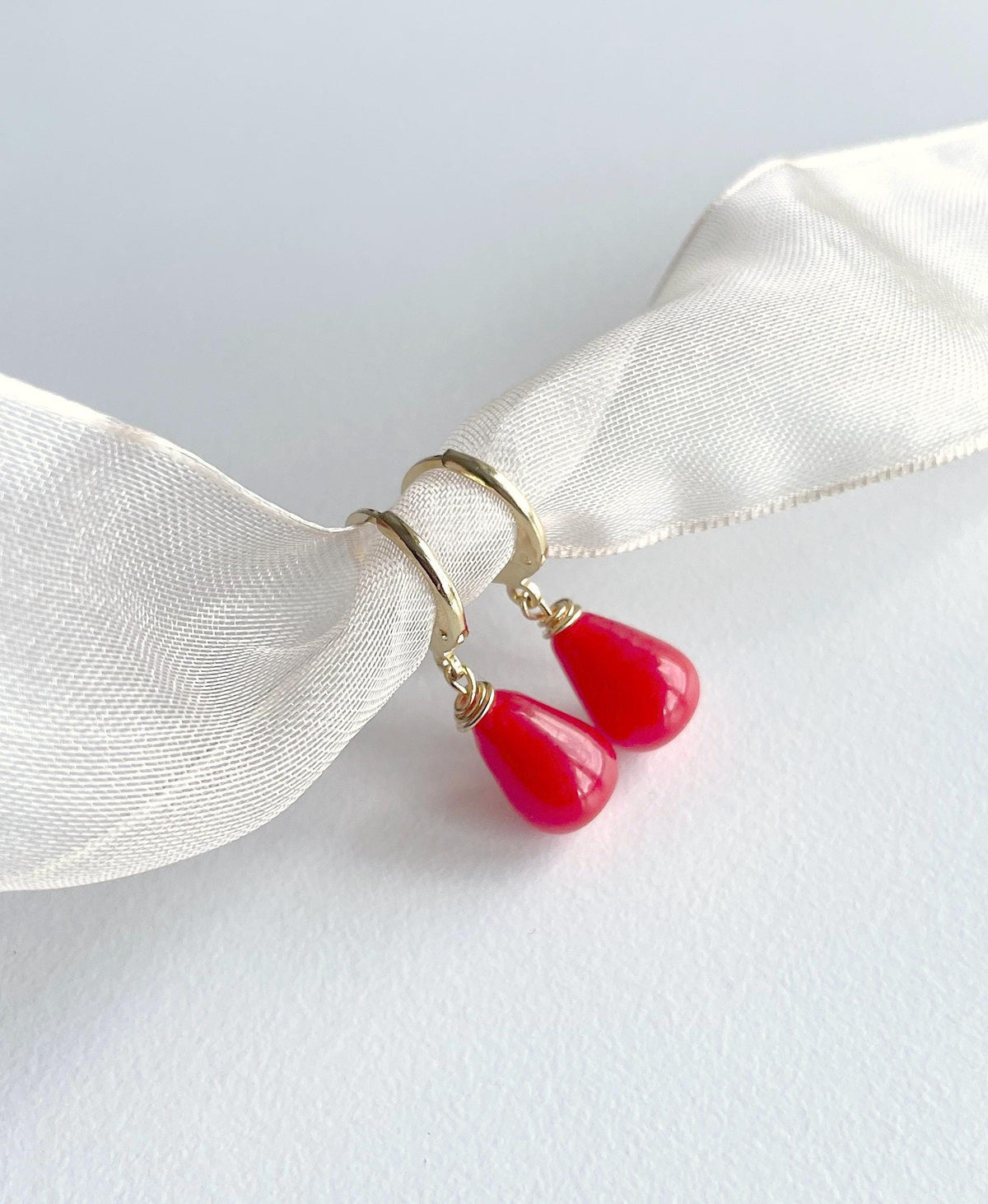 A pair of gold hoop earrings with bright red teardrop-shaped drop charms, displayed on a white satin ribbon against a light gray background.