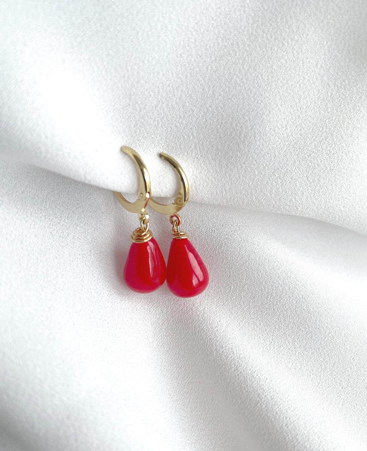 A pair of gold huggie hoop earrings with dangling red teardrop beads, displayed on a soft, white textured fabric background.