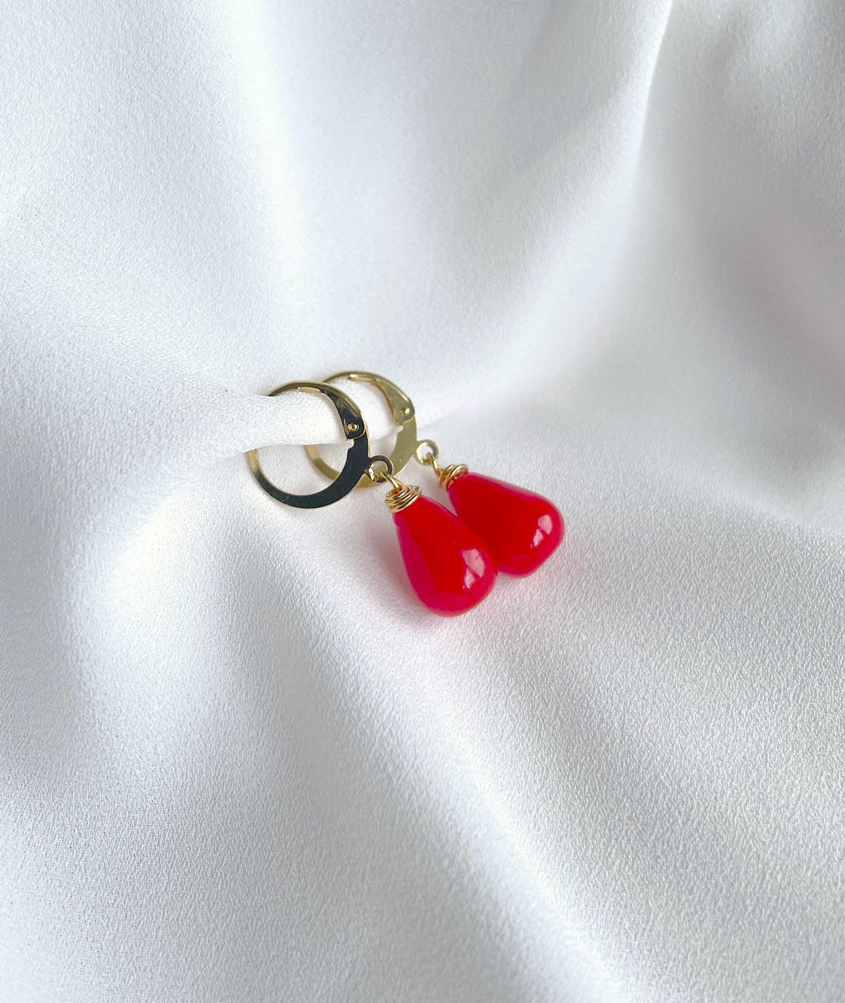 A pair of gold hoop earrings with dangling red teardrop charms, displayed on a draped white fabric background.