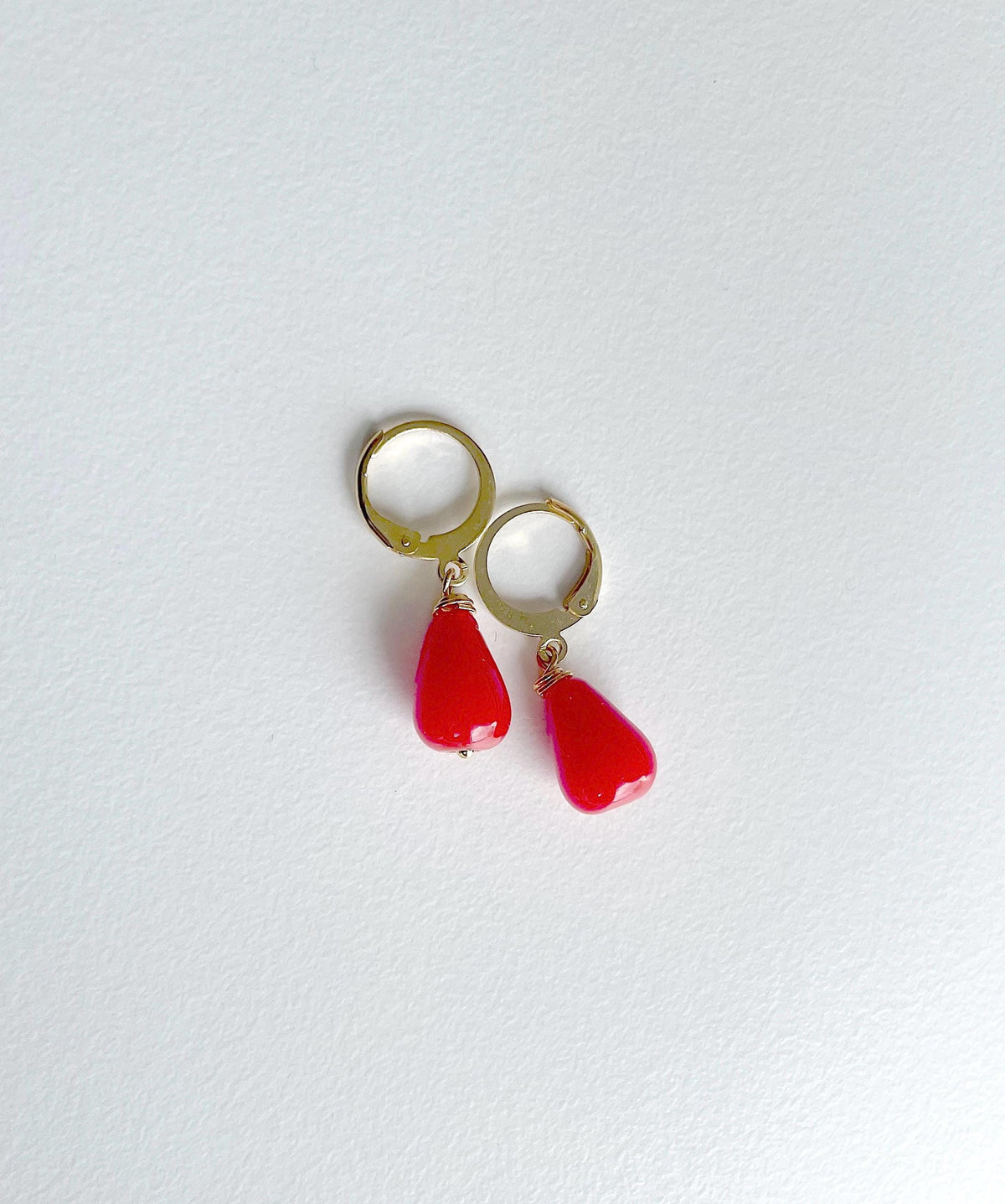 A pair of gold hoop earrings with dangling red teardrop-shaped beads, displayed on a clean, light off-white textured surface.