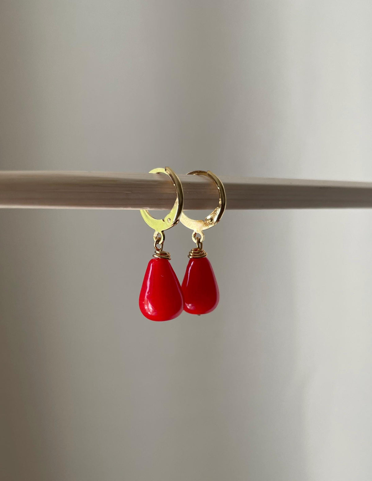 A pair of gold hoop earrings with bright red teardrop-shaped drop charms, hanging from a thin wooden rod against a soft neutral background.