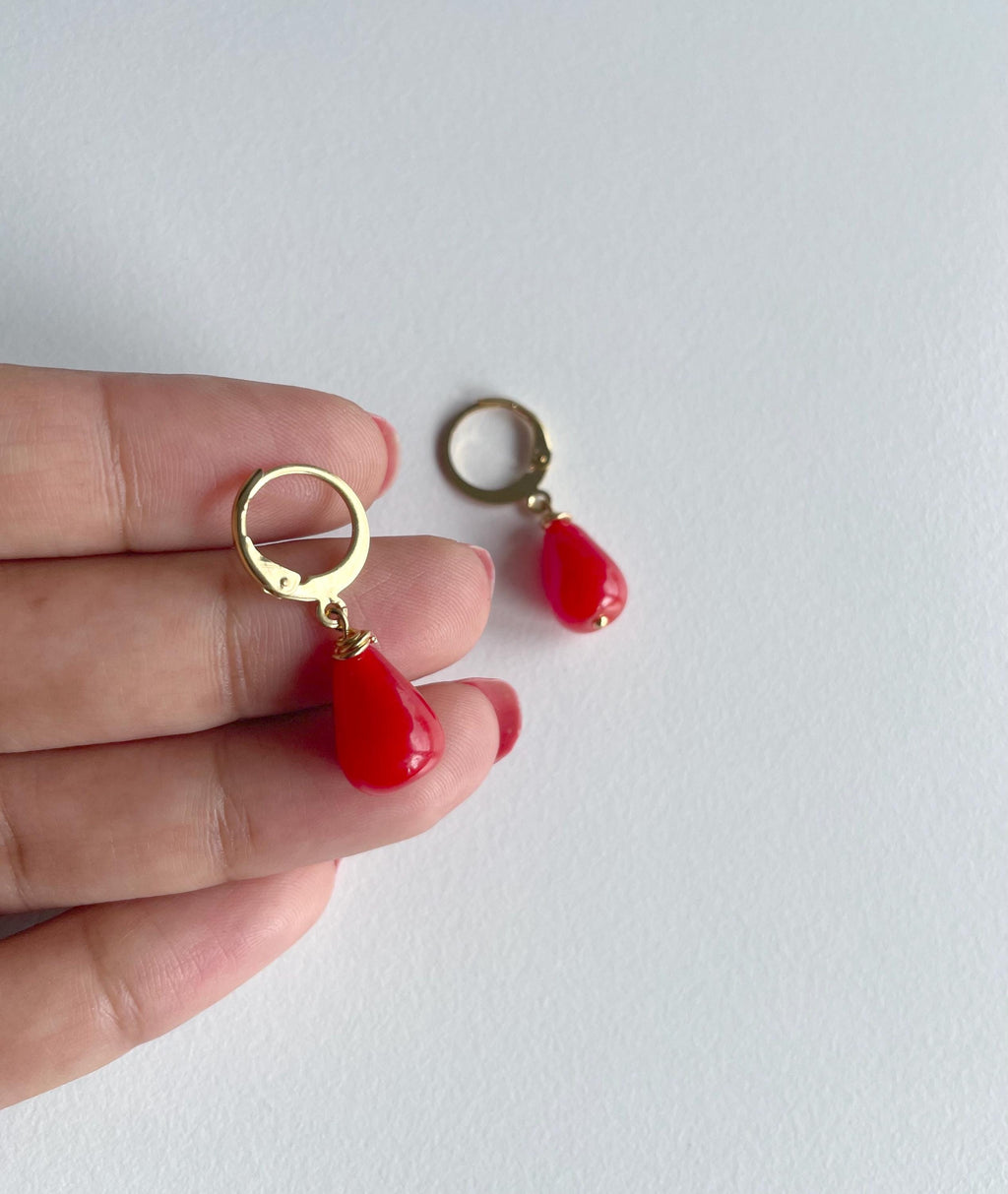 A pair of gold hoop earrings with red teardrop-shaped drop charms, shown with one earring held on a person's fingers and the other placed on a light gray surface.