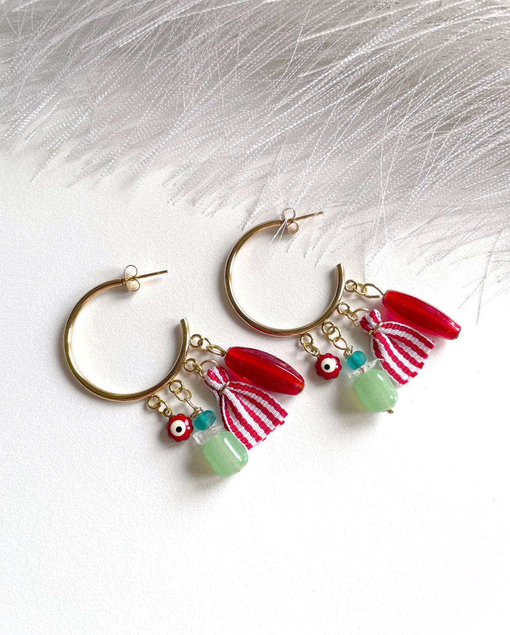A pair of gold-tone hoop earrings with dangling charms, including red beads, a red-and-white striped ribbon, a small evil eye bead, and a light green bead, displayed on a white textured surface with white feathers.