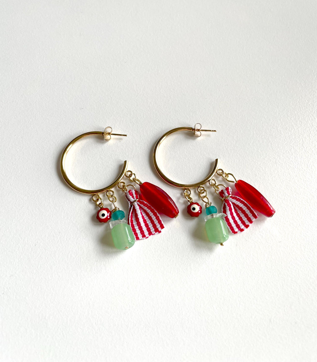 A pair of gold-tone hoop earrings with dangling charms, including red and white striped tassels, red curved beads, and light green beads with evil eye details, displayed on a white background.