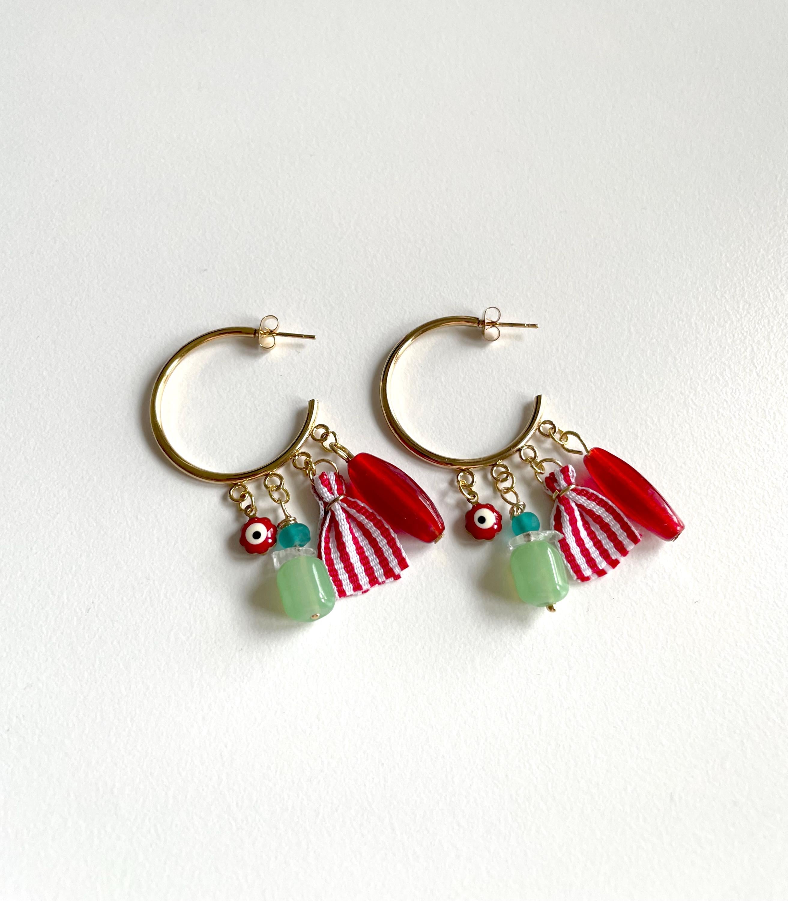 A pair of gold-tone hoop earrings with dangling charms, including red and white striped tassels, red curved beads, and light green beads with evil eye details, displayed on a white background.